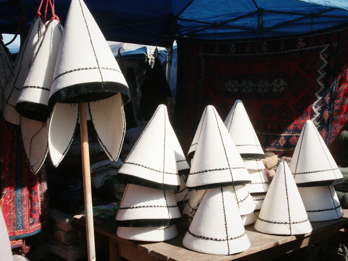 Tall pointed white felt hats with black embroidered seams and black velvet brims stacked on a wooden table and hanging from a pole under a blue awning, with a red patterned carpet behind and a man looking on at the right edge