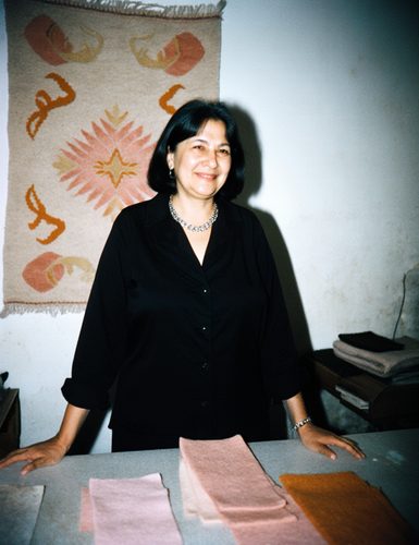 A smiling woman in a black blouse at a table of felt swatches in front of a pale felt rug with ram's-horn patterns