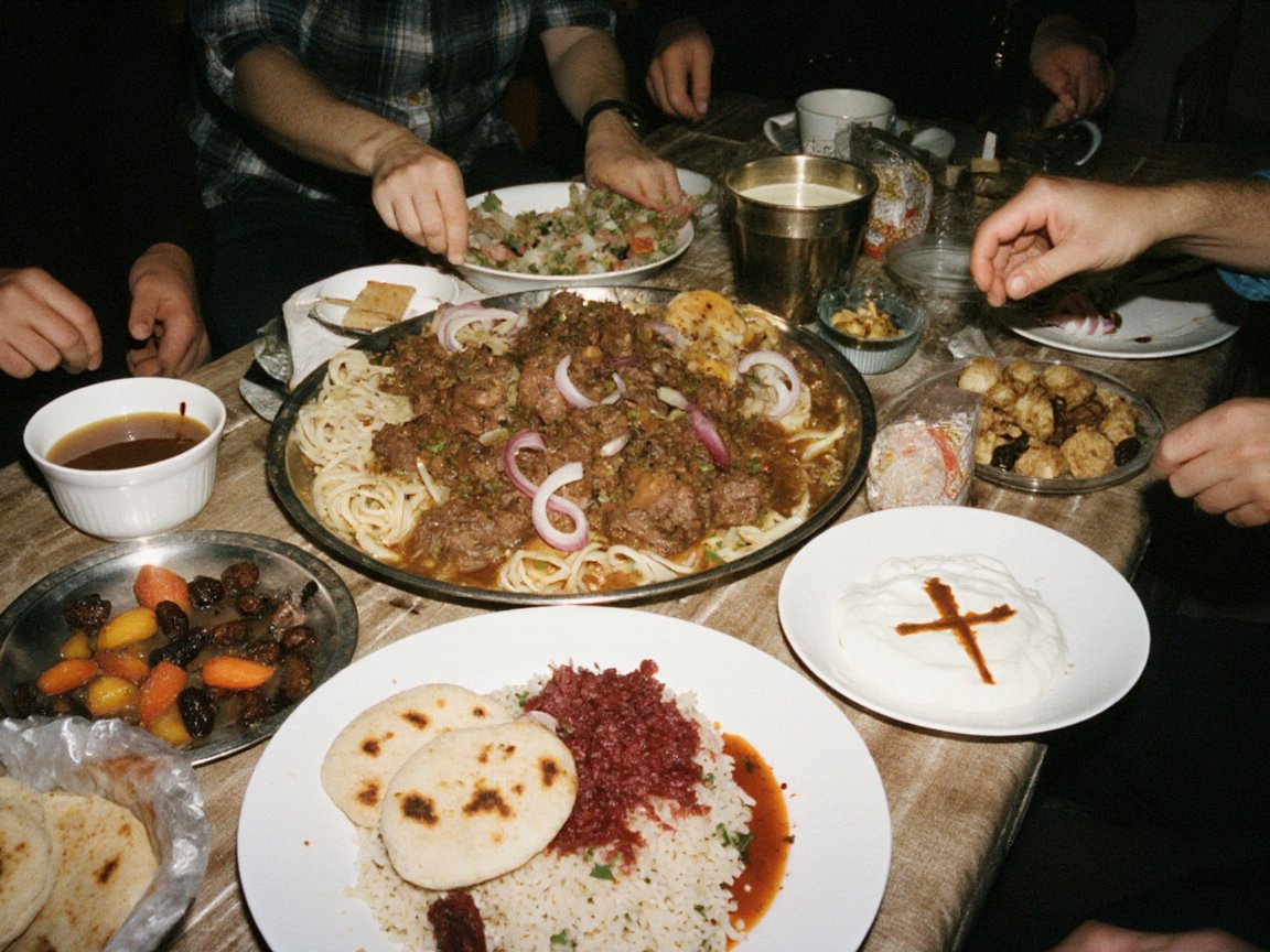 Flash photo of a crowded table: a platter of noodles with stewed meat and onion, rice topped with red beetroot and flatbreads, yoghurt marked with a cross, soups and sweets, hands reaching in