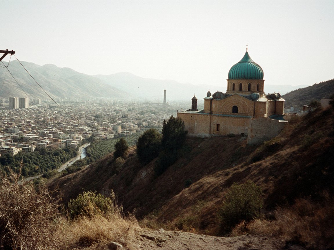 Yellow-brick church with a turquoise dome on a dry hill spur at the right, above a river and a large city filling a valley among hazy mountains; a wooden electricity pole and dry grass in the foreground
