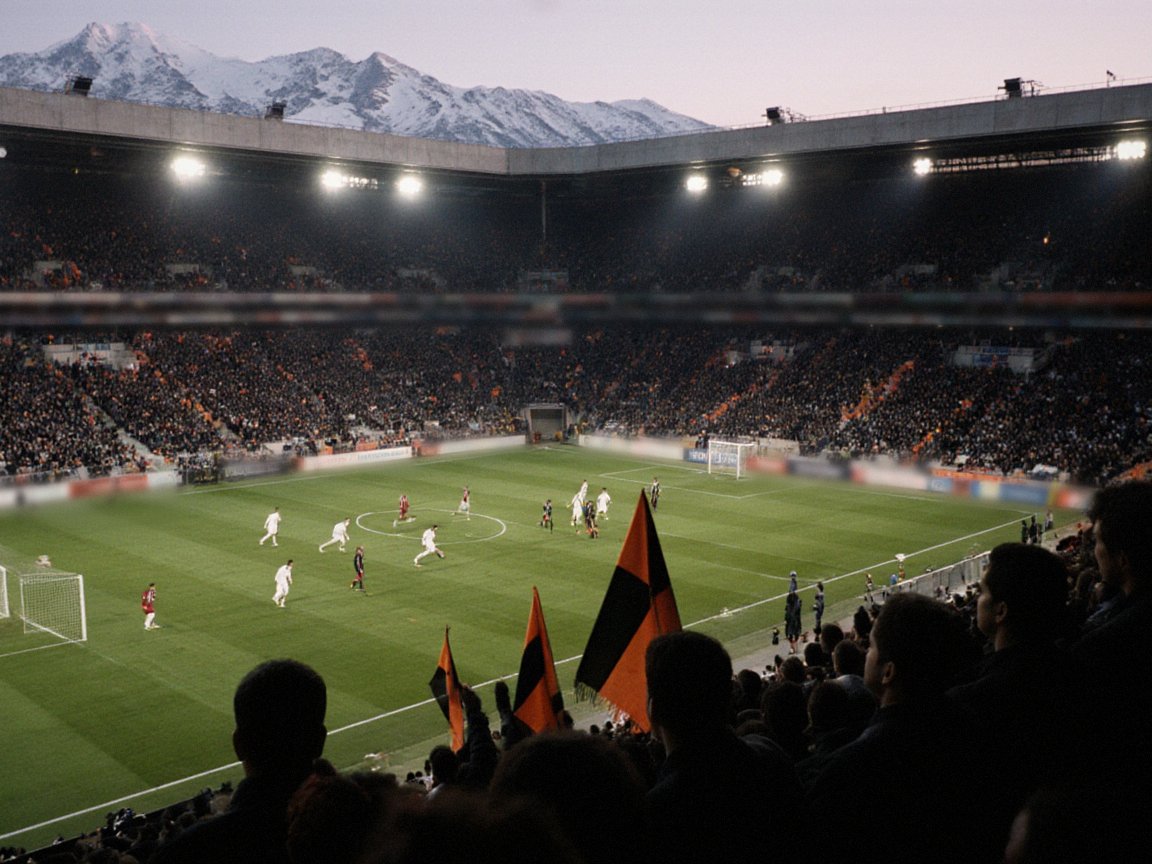 An evening match under floodlights seen from high in a full stand, with players in white and in crimson on the pitch, black-and-orange flags waved in the foreground and snowy mountains above the roof