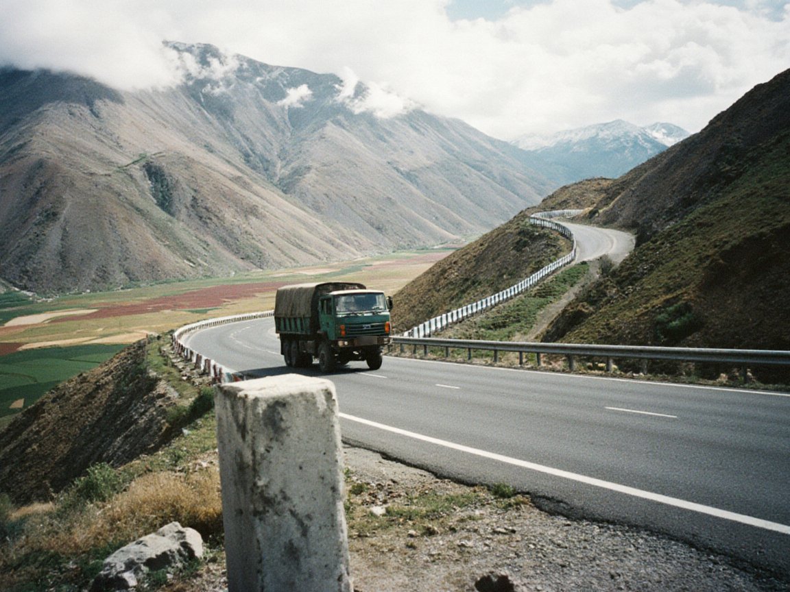 A green lorry rounds a bend on a mountain road with crash barriers above a broad valley of patchwork fields, with a concrete marker post in the foreground and clouds on the ridges