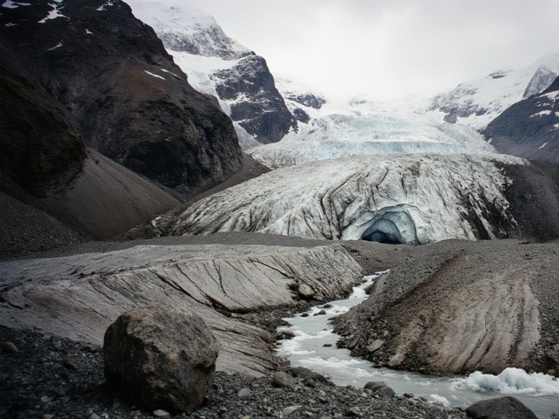 The grey, crevassed snout of a valley glacier ending in an ice cave, with a meltwater stream running out across bare moraine and a large boulder in the foreground, below snowy peaks under cloud