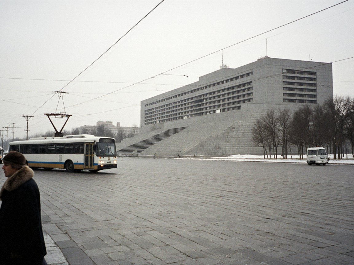 Woman in a fur-collared coat at the edge of a vast paved square in winter; a trolleybus under overhead wires and a large stepped grey concrete government building set at an angle on the right