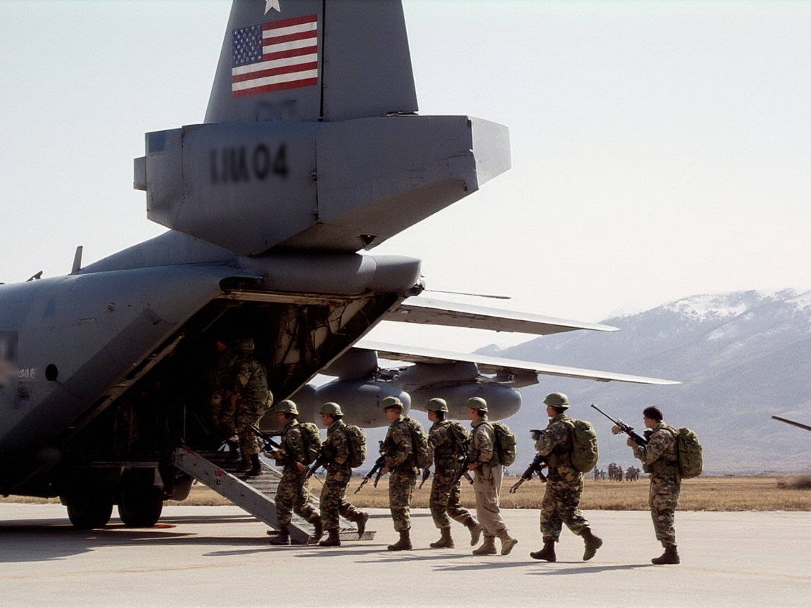 Soldiers in camouflage with packs and rifles file up the rear ramp of a grey four-engined transport aircraft with a US flag on its tail, a helicopter on the grass beyond and snow-streaked mountains behind