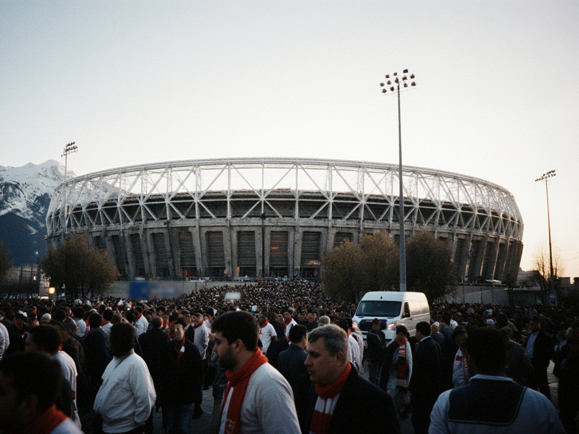 A large crowd of fans in red-and-white scarves walking toward a round stadium with a white steel roof frame over a concrete bowl, with floodlights and a snowy mountain at dusk